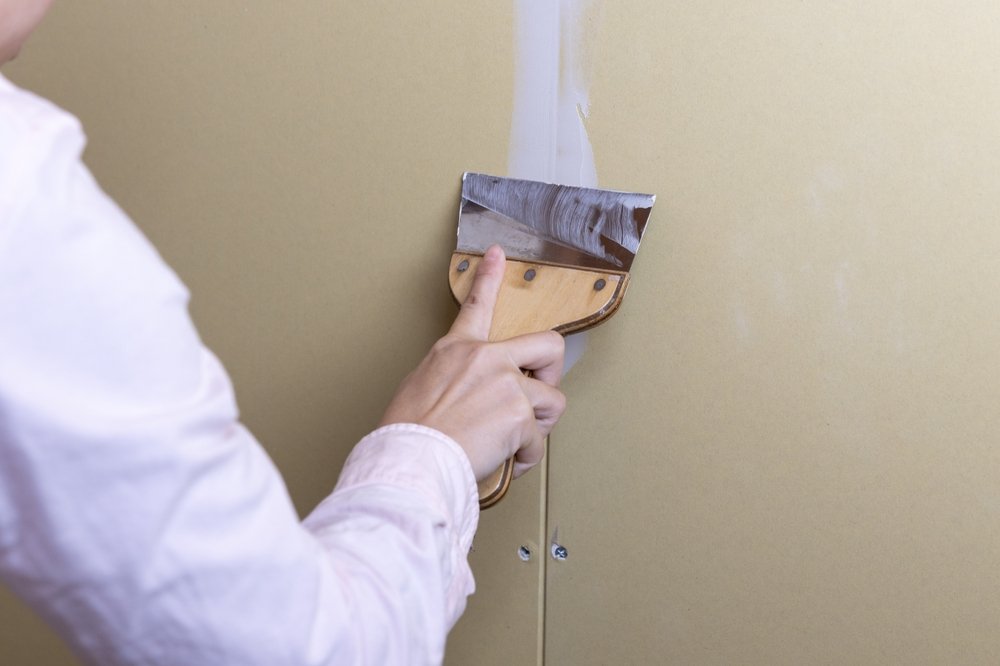 Een afbeelding van een persoon die een muur behangklaar maakt door het vullen van scheuren en het gladmaken van het oppervlak, als voorbereiding voor het aanbrengen van behang.