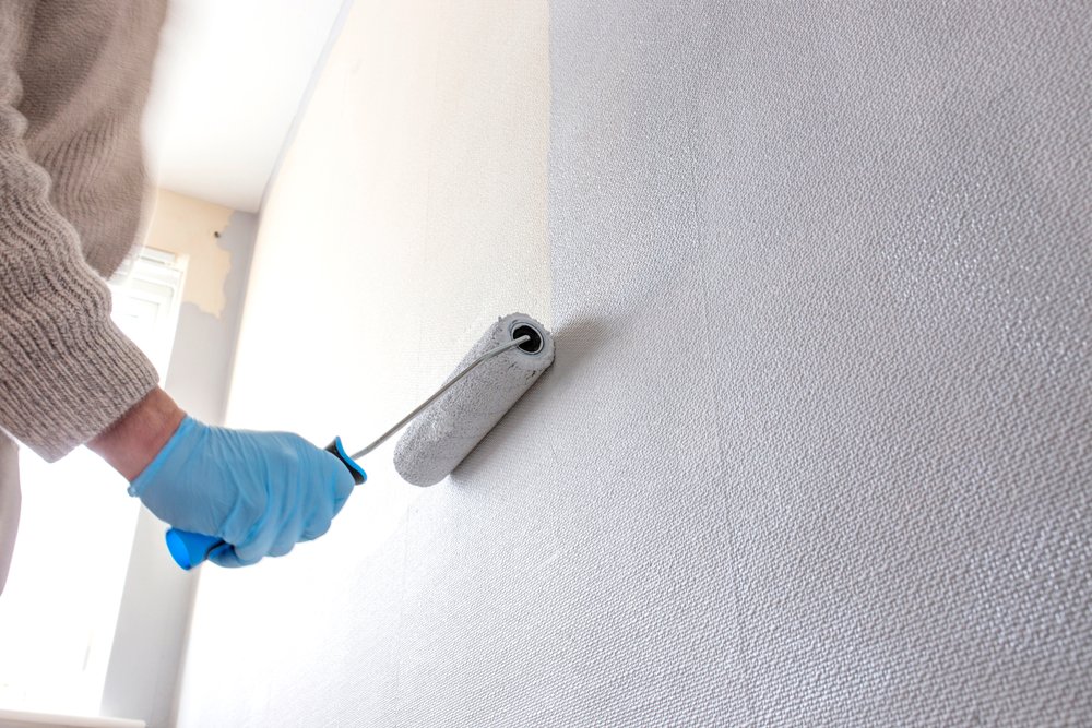 Een afbeelding van een hand die een roller gebruikt om verf aan te brengen op behang aan de muur, als onderdeel van het proces van behang schilderen voor een vernieuwde interieurlook.